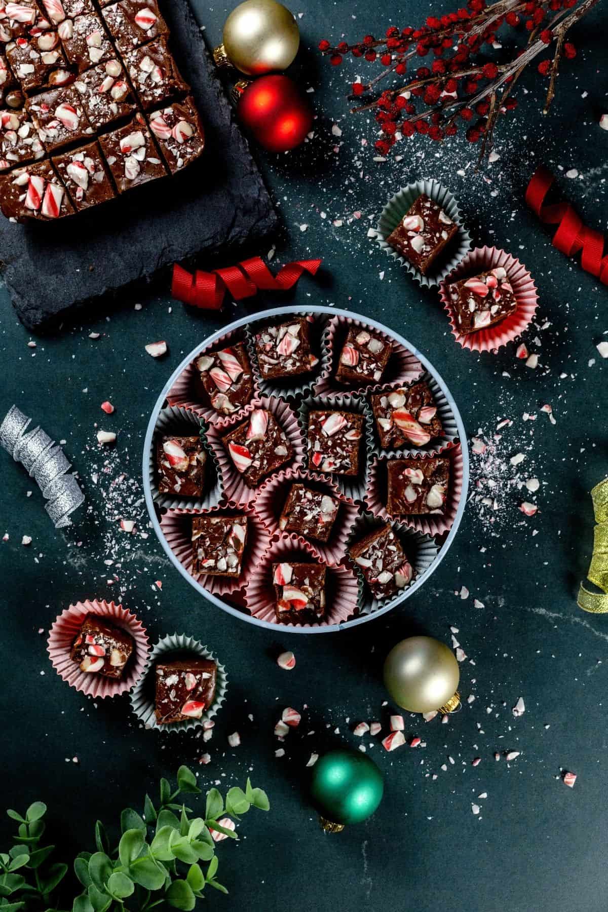 a round tin is filled with pieces of Christmas fudge covered in crushed candy canes. more pieces are scattered around the table, along with ribbons, more candy canes, and a few Christmas ornaments.