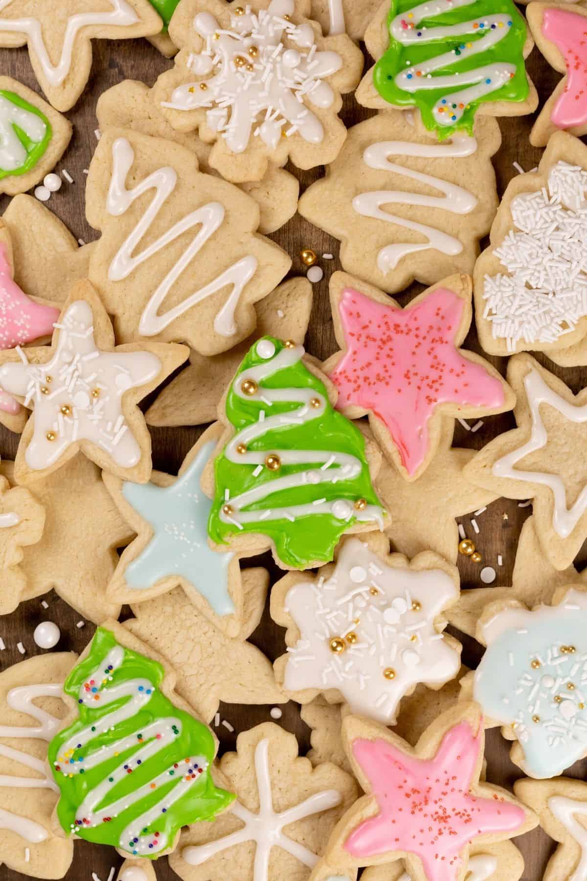 a large pile of Christmas sugar cookies stacked on top of each other in various shapes like trees, stars, and snowflakes. some are covered in pastel frosting with sprinkles and some are plain.