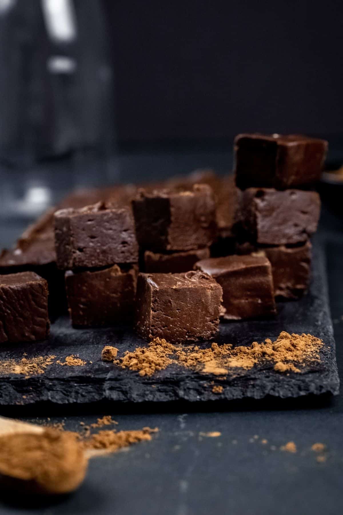 stacks of vegan chocolate fudge on a dark serving tray. the fudge is cut into little cubes. fresh cocoa powder is sprinkled around the fudge.
