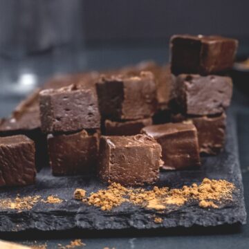 stacks of dairy free fudge cut into cubes on a dark serving tray. fresh cocoa powder surrounds the fudge.