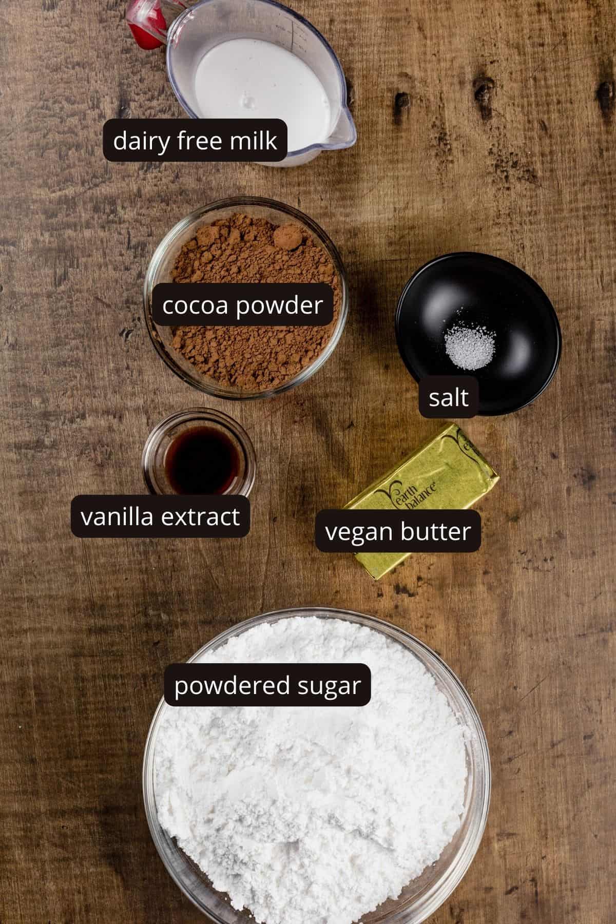 ingredients for fudge in various glass bowls resting on a wood table. black and white labels have been added to name each ingredient.