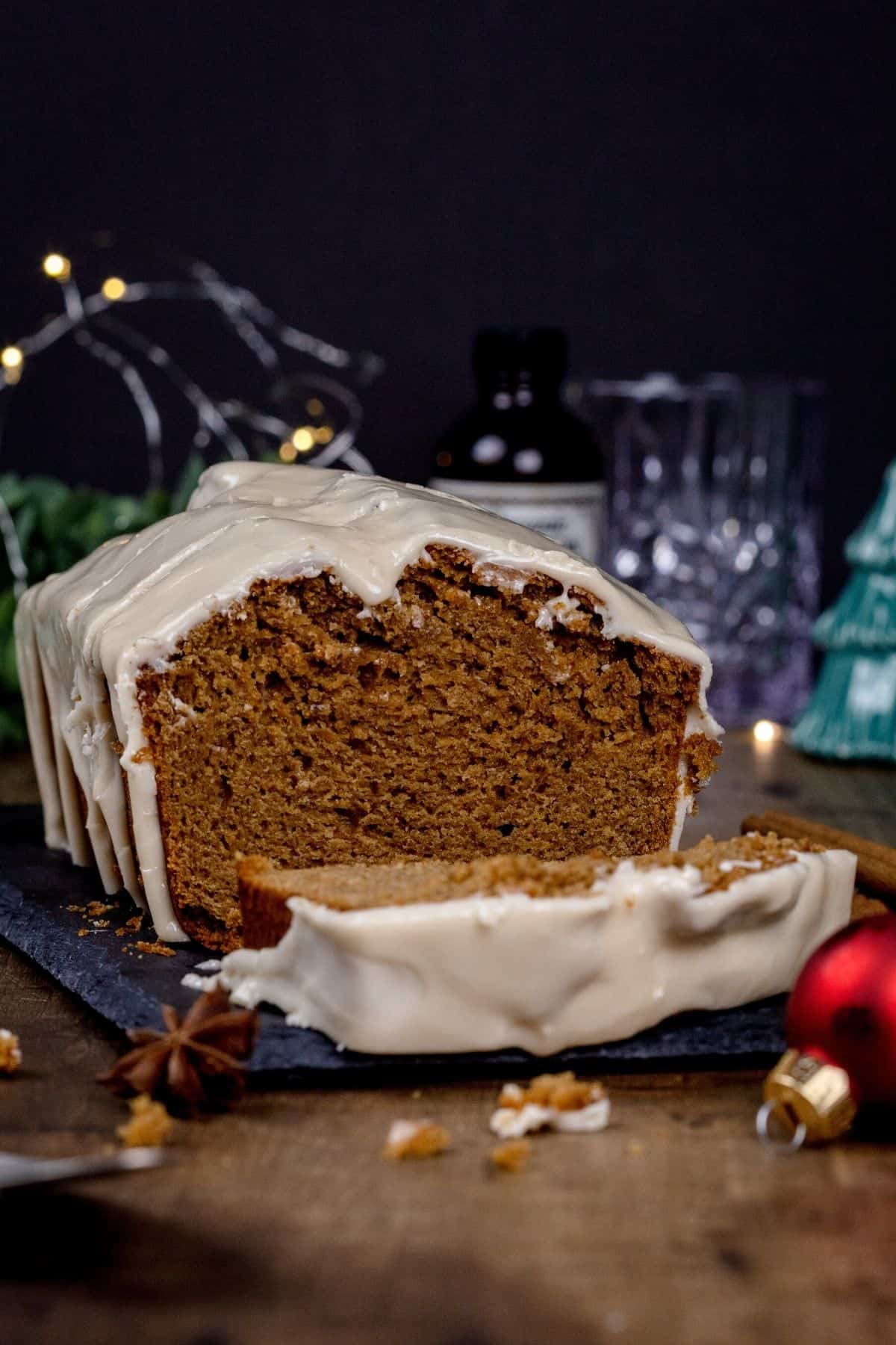 ¾ view of a sliced loaf cake with a slice in front. crumbs surround the cake, as well as spices and Christmas decor. lights are blurred in the background.
