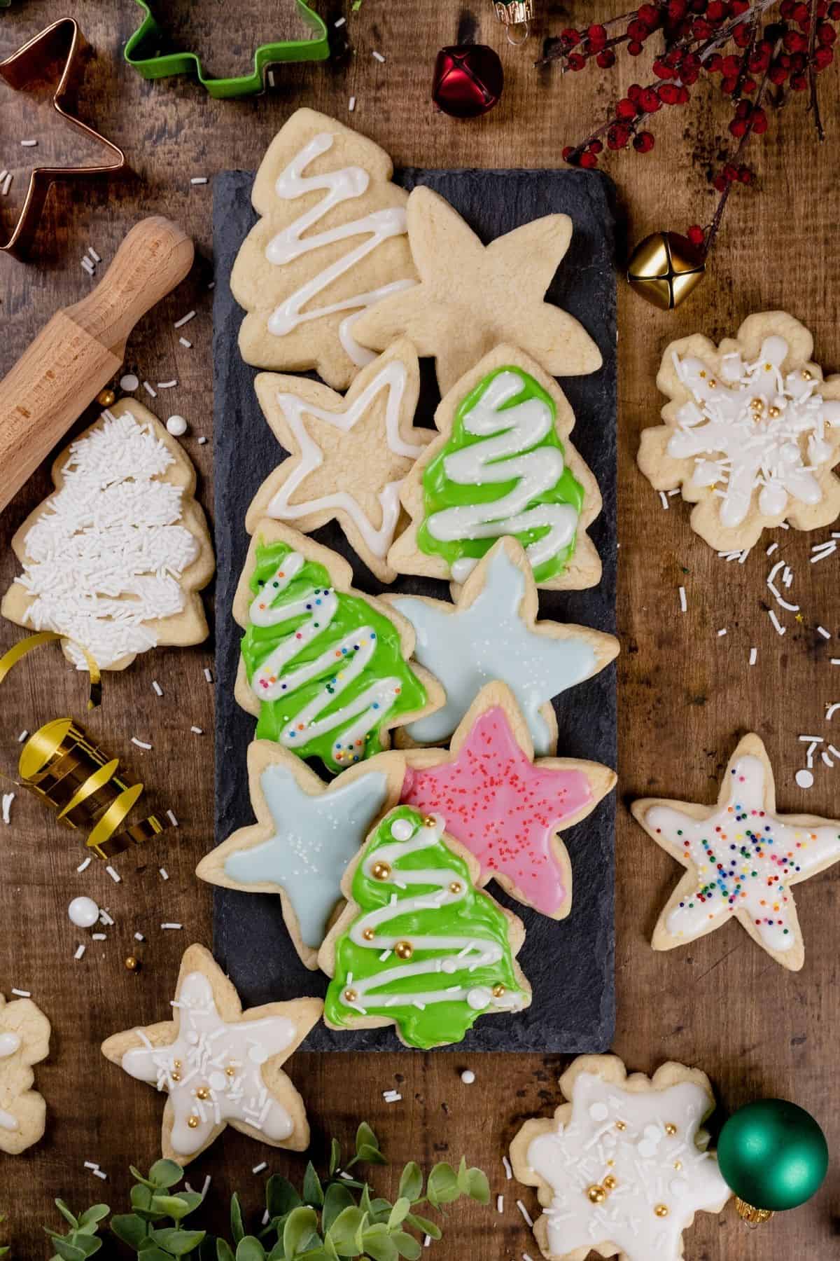 black plate filled with Christmas sugar cookies in various shapes like trees and stars. some are covered in icing and sprinkles and some are plain. more cookies, sprinkles, ribbons, and various holiday themed items surround the plate making for a very full picture.