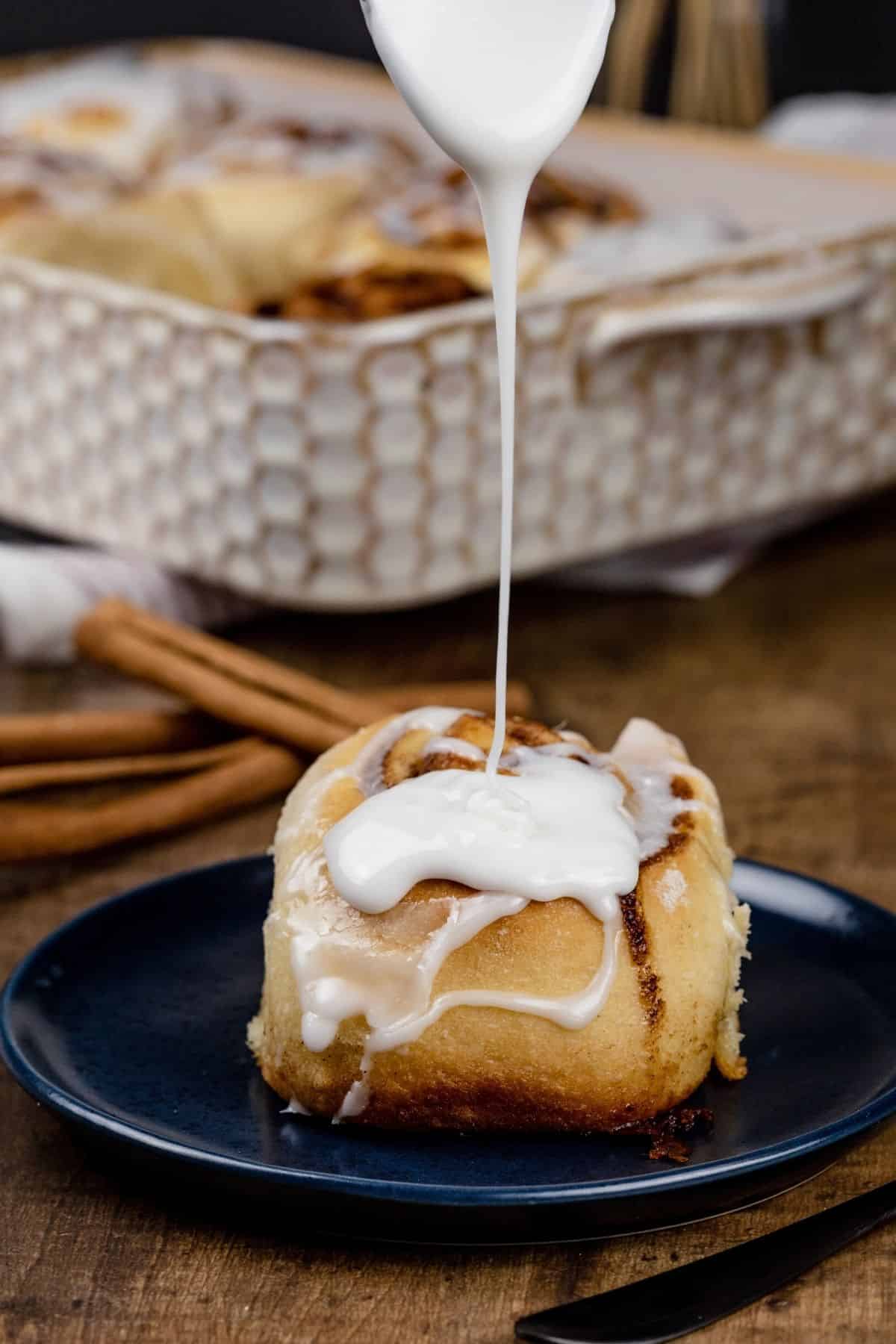 a single cinnamon roll is on a dark blue plate in front of a dish filled with the other rolls. a spoon is drizzling thick white icing on top of the roll.