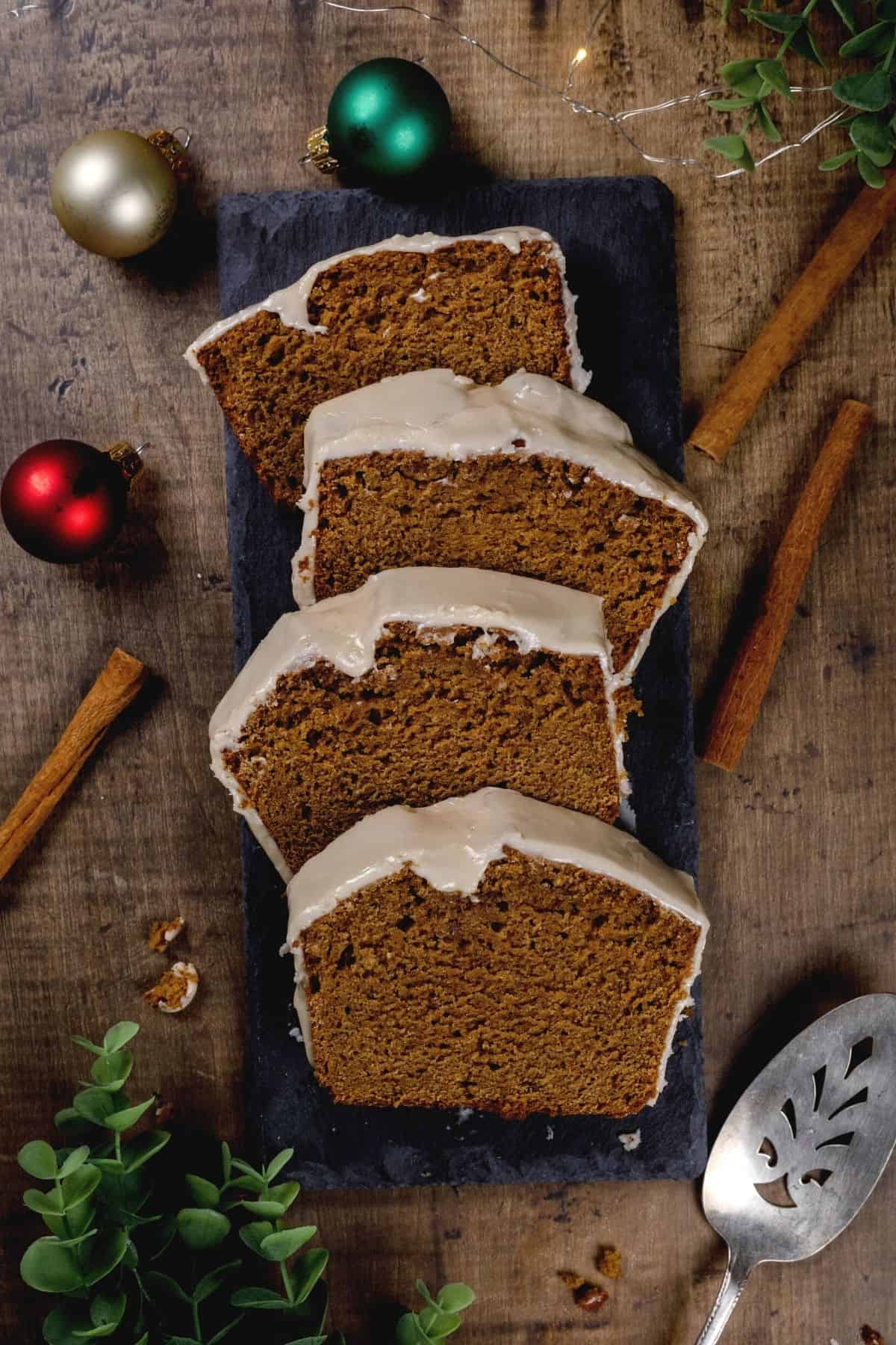 4 slices of gingerbread cake on a black slate. a serving spoon is seen. cinnamon sticks and Christmas ornaments surround the cake.