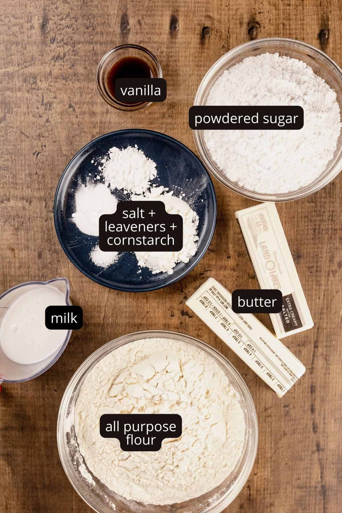 ingredients for cookies in various glass bowls on a wood tabletop. black and white labels have been added to name each ingredient.