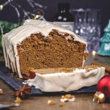 iced gingerbread cake on a black serving tray. a slice is blurred in the front of the loaf. lights and Christmas themed decor is blurred around the cake. spices and crumbs are also seen.