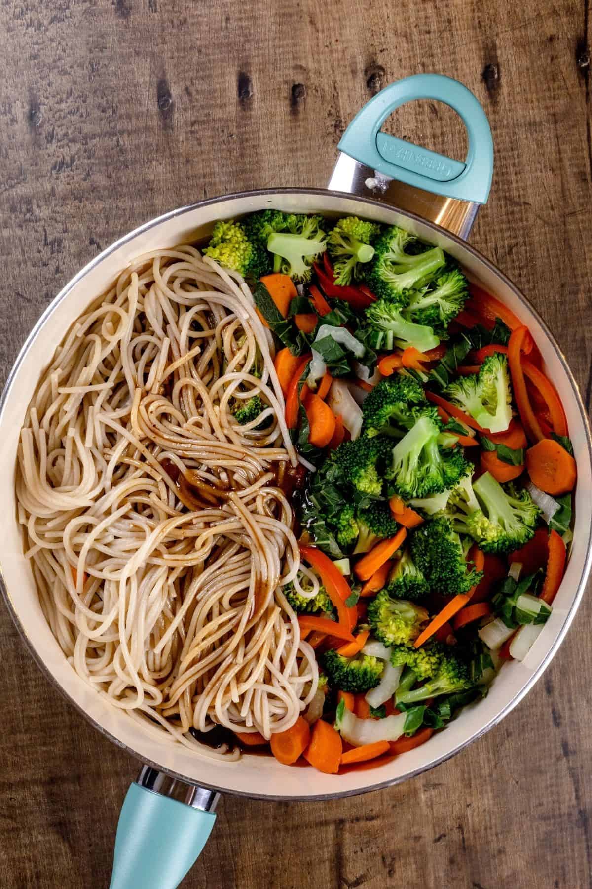 A blue and white fry pan is half filled with cooked veggies and half filled with brown rice noodles. The sauce is poured over top. The pan is on a wood tabletop.