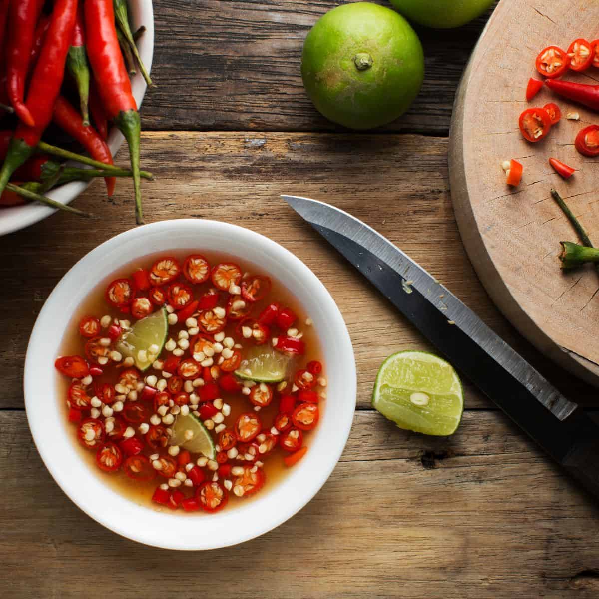 A small white bowl is filled with fish sauce, chilis, and pieces of lime. Chilis and limes and seen next to a cutting board with a knife.