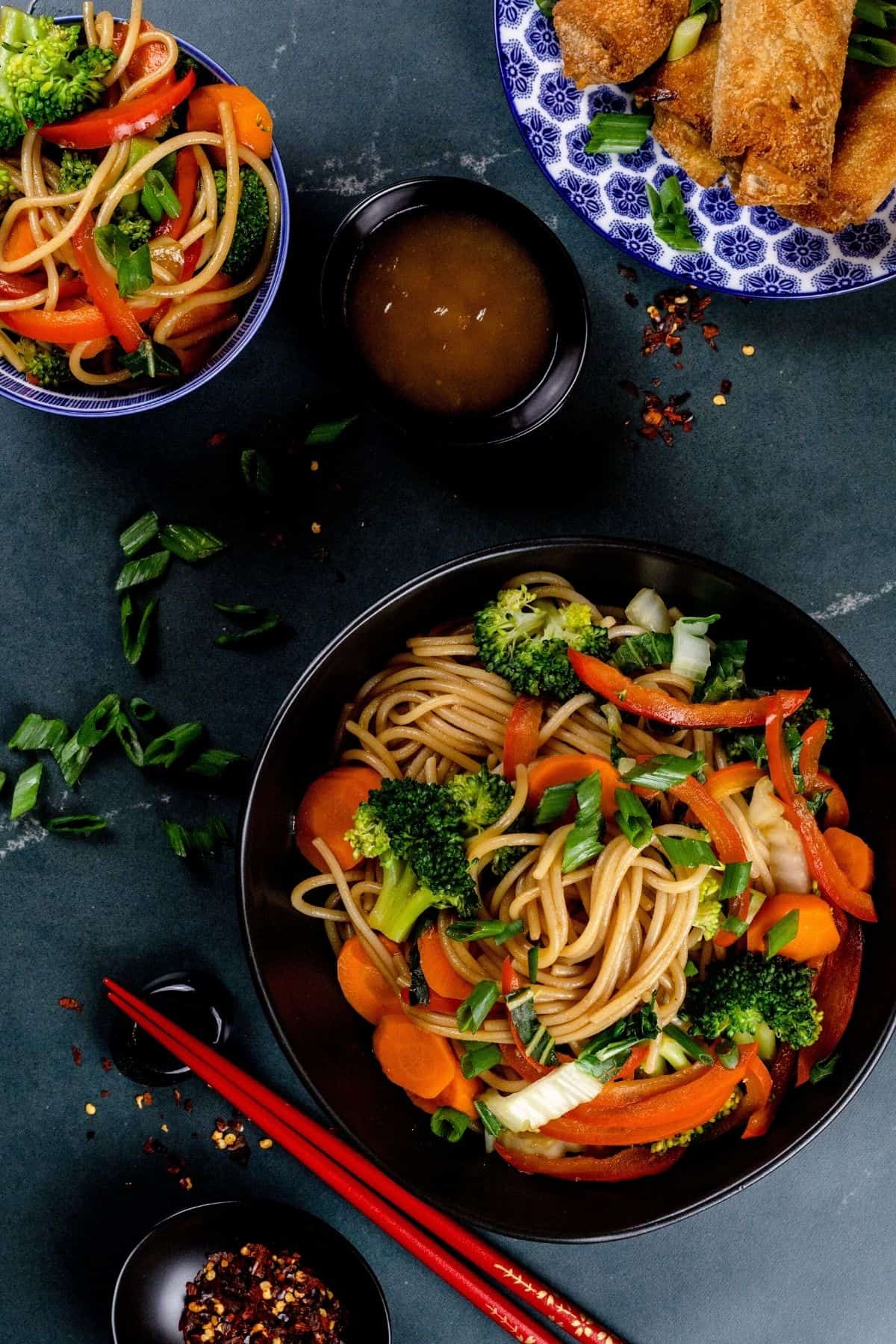Many bowls of lo mein with a plate of egg rolls. Tiny bowls filled with red pepper flakes and dipping sauce are also seen. Red chopsticks are next to the bowls. Sliced green onions are sprinkled around.