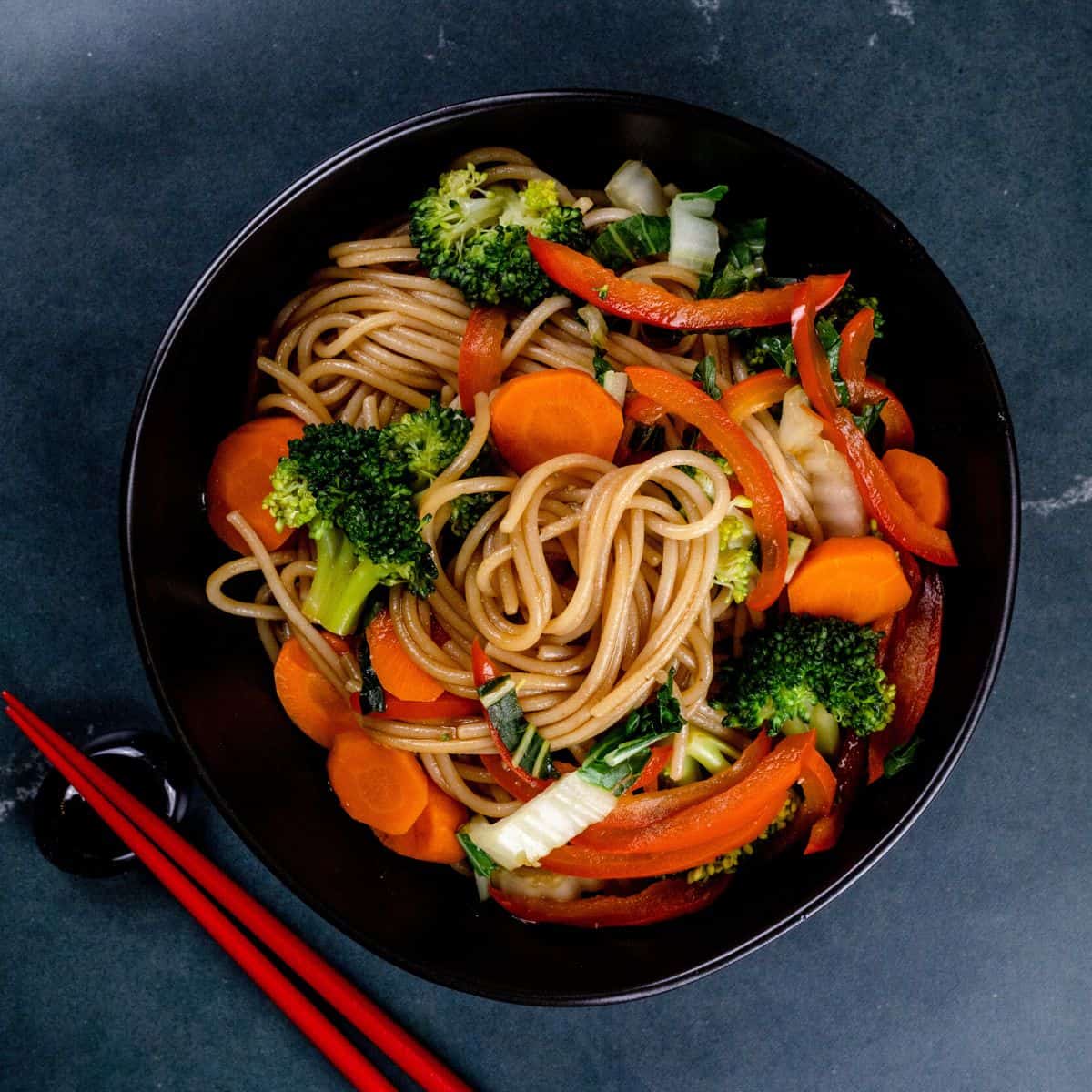 A dark bowl is filled with veggie lo mein. The noodles are swirled in the bowl with lots of veggies in them. A pair of red chopsticks are next to the bowl. It is resting on a dark marble surface.