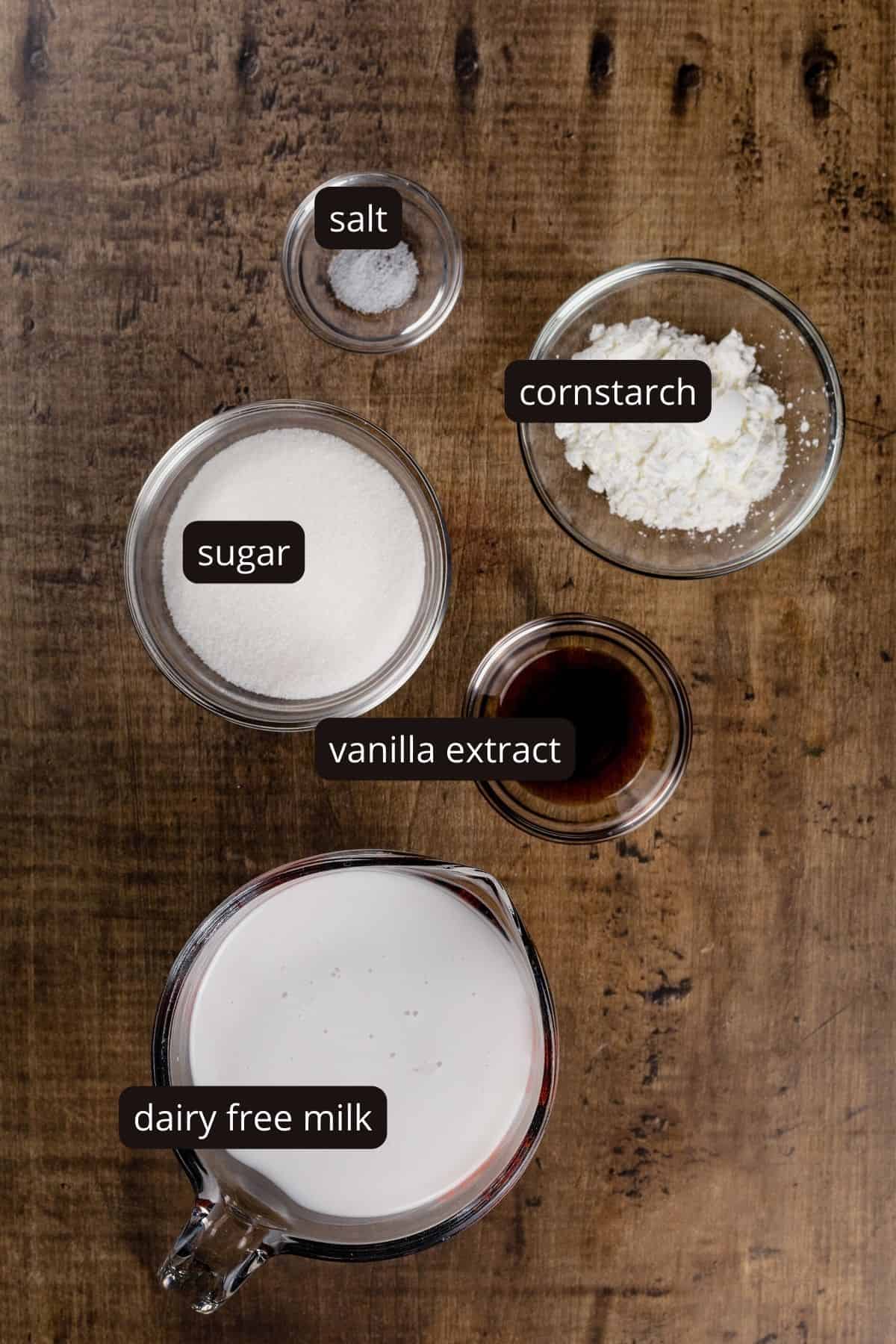 Ingredients for pudding in various glass bowls of different sizes rest on a wood table. Black and white labels have been added to name each ingredient.