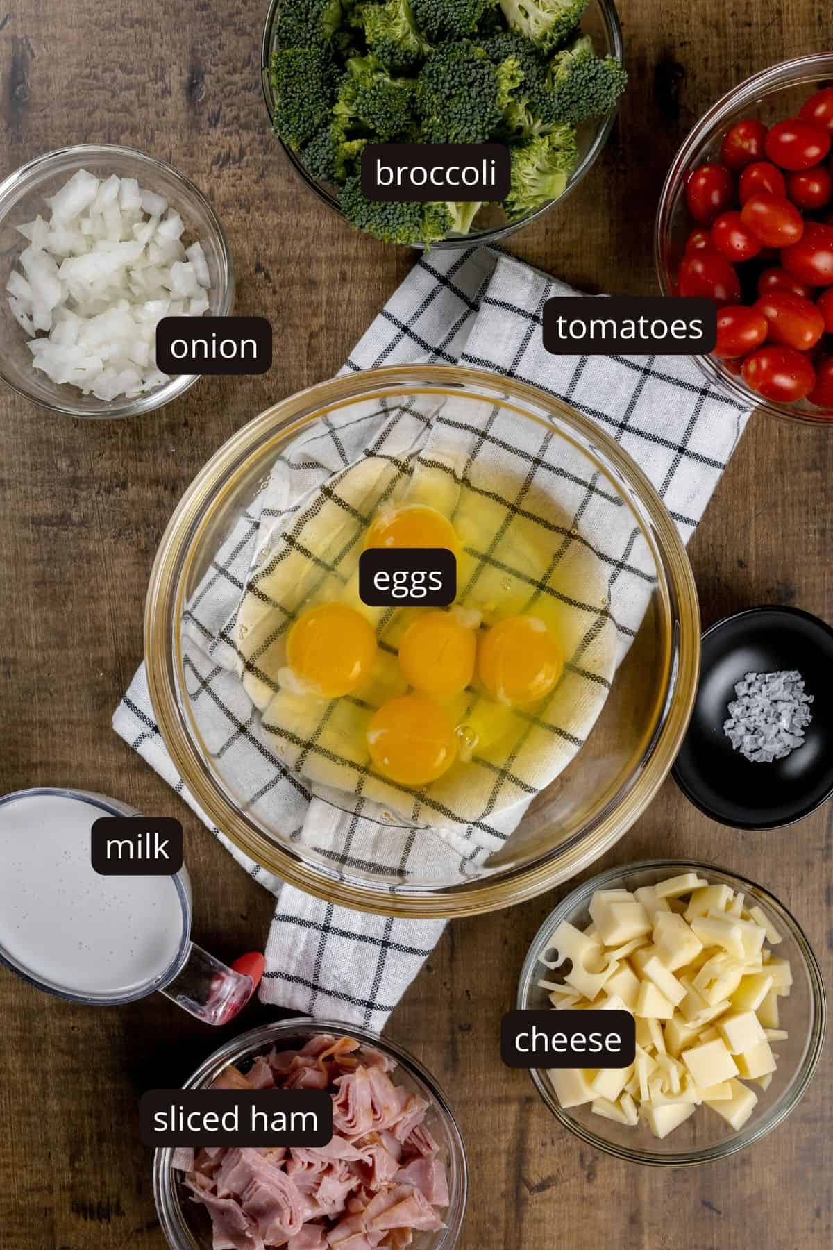 The different fillings for the quiche are in various glass bowls on a wood tabletop surrounding a large bowl willed with cracked eggs. Black and white labels have been added to name each ingredient.