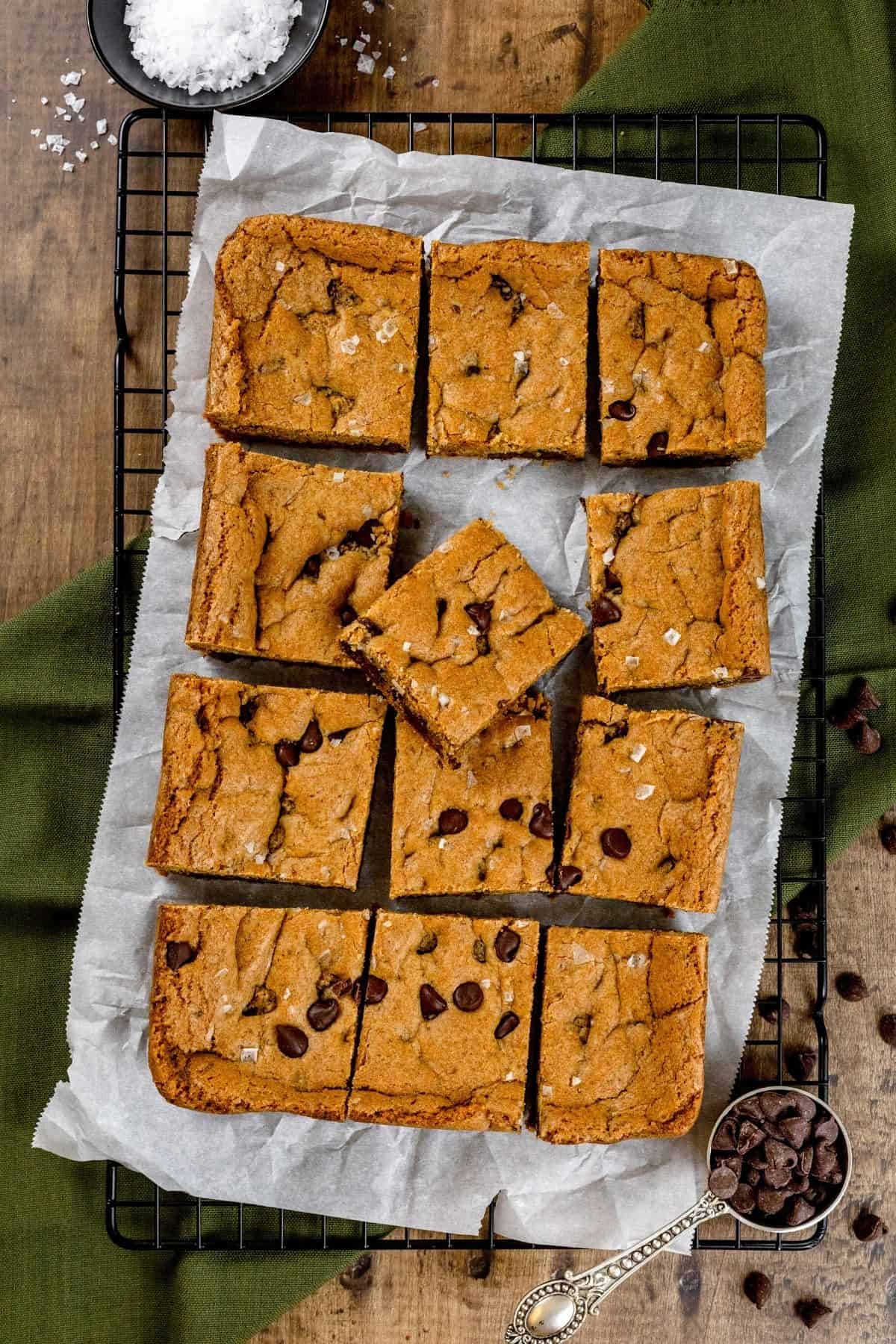 12 slices of chocolate chip bar cookies with one cookie resting on a tilt on crinkled parchment paper on a wire cooling rack with a green towel, a bowl filled with flaky sea salt, and a metal scoop filled with chocolate chips. All on a wood tabletop.
