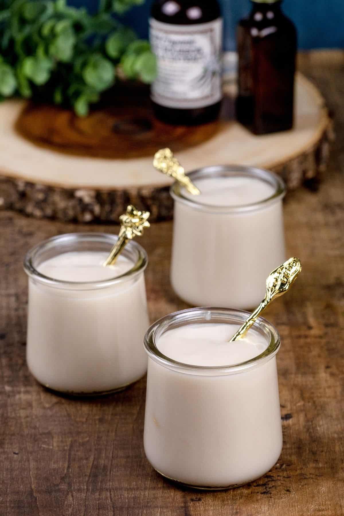 3 vanilla glass pudding cups with gold spoons in each of them. Vanilla bottles are blurred in the background on a wood tray.