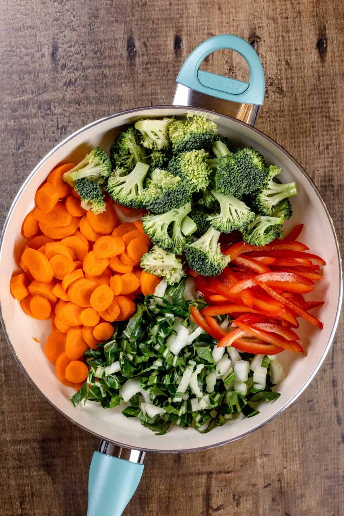 Uncooked veggies like broccoli, carrots, and bell peppers in a blue and white skillet on a wood table.
