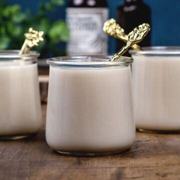 3 glass cups filled with vegan vanilla pudding. Each has a small gold spoon in the pudding cup. Bottles of vanilla are blurred in the background.