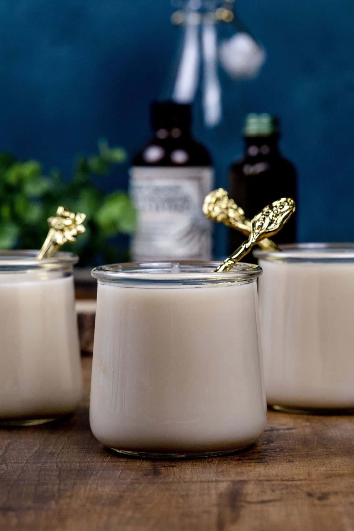 3 glass cups filled with vegan vanilla pudding. Each has a tiny gold spoon in the cup. Bottles of vanilla are blurred in the background.