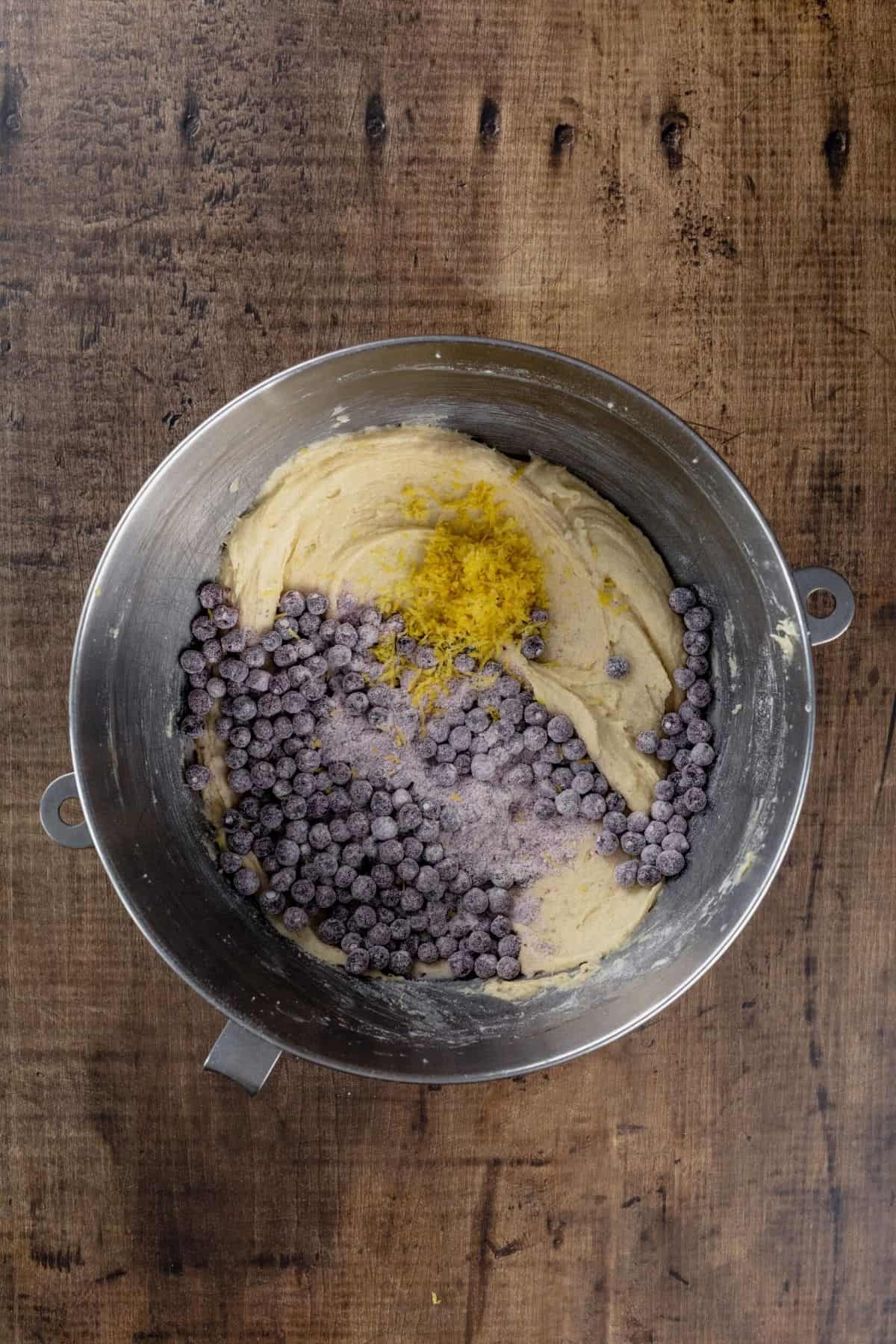 A silver bowl of muffin batter with many blueberries being added.