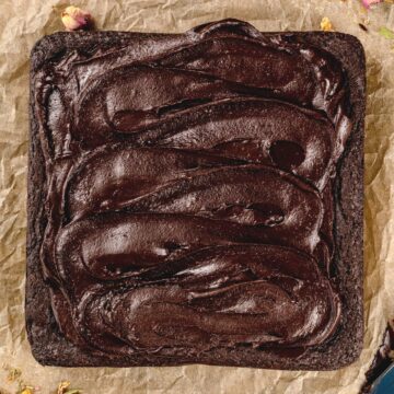 A batch of brownies on crinkled brown parchment paper with lots of creamy chocolate frosting on top of the brownies.