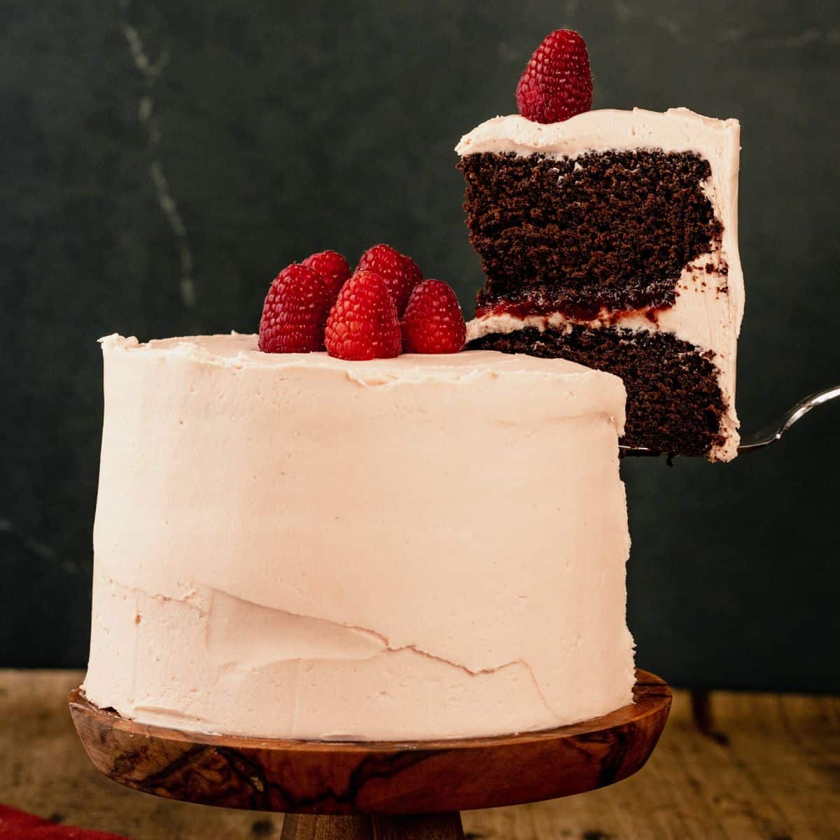 A chocolate raspberry cake covered in pink frosting on a wood cake stand with a slice of cake being lifted out of the cake.