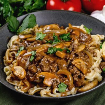 Dark bowl filled with noodles and dairy free beef stroganoff. Fresh parsley is sprinkled on top. More fresh parsley, tomatoes, and mushrooms are blurred in the background.