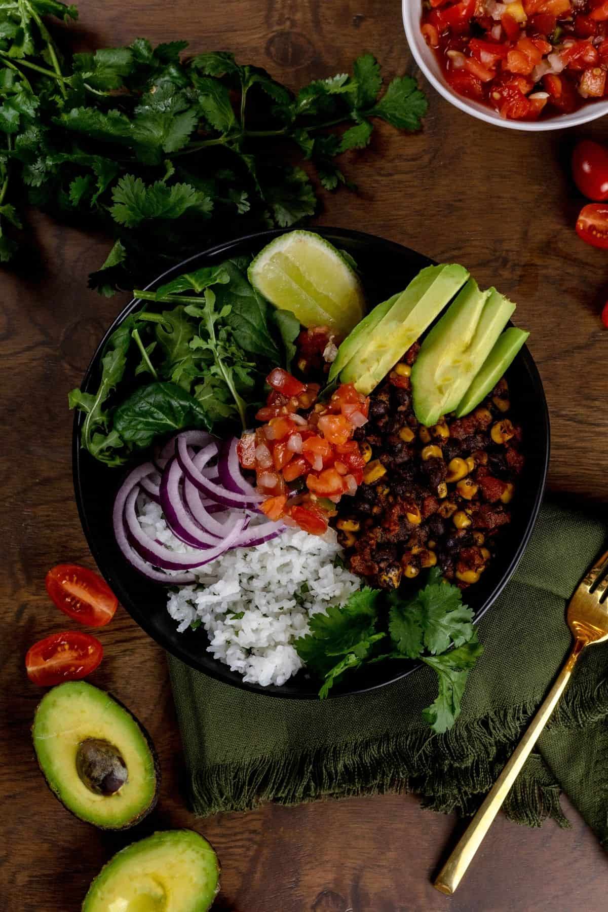 A look down at a dark bowl on a wood table filled with a veggie burrito bowl. Things like rice, beans, corn, salsa, onion, greens, avocado, and more are piled high in the bowl with extras scattered around the bowl on the tabletop. A green napkin and gold fork are in the lower right corner.