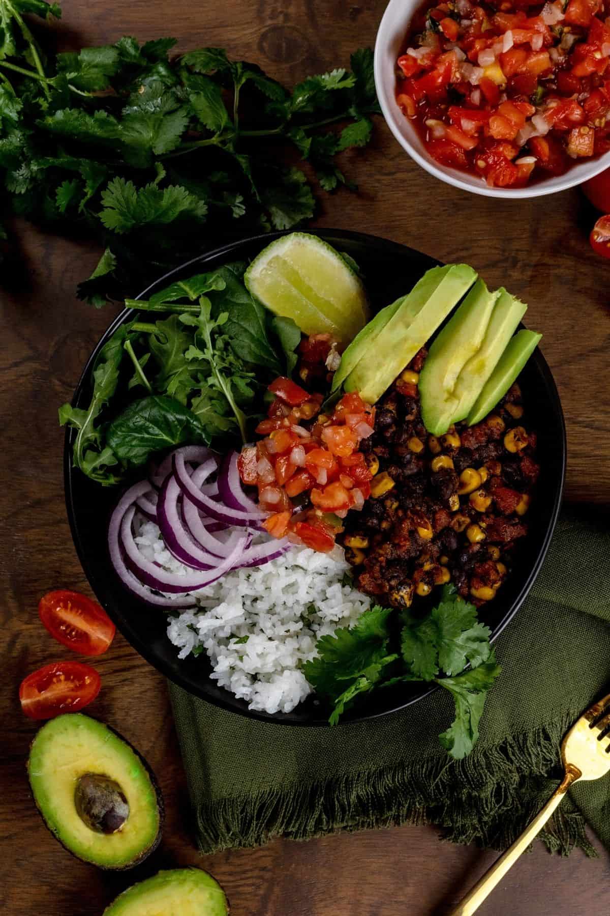 A closer look at a dark bowl on a wood table filled with a veggie burrito bowl with rice, beans, avocado, salsa, and more. Extra ingredients are around the bowl. A green napkin and gold fork are in the lower right corner.