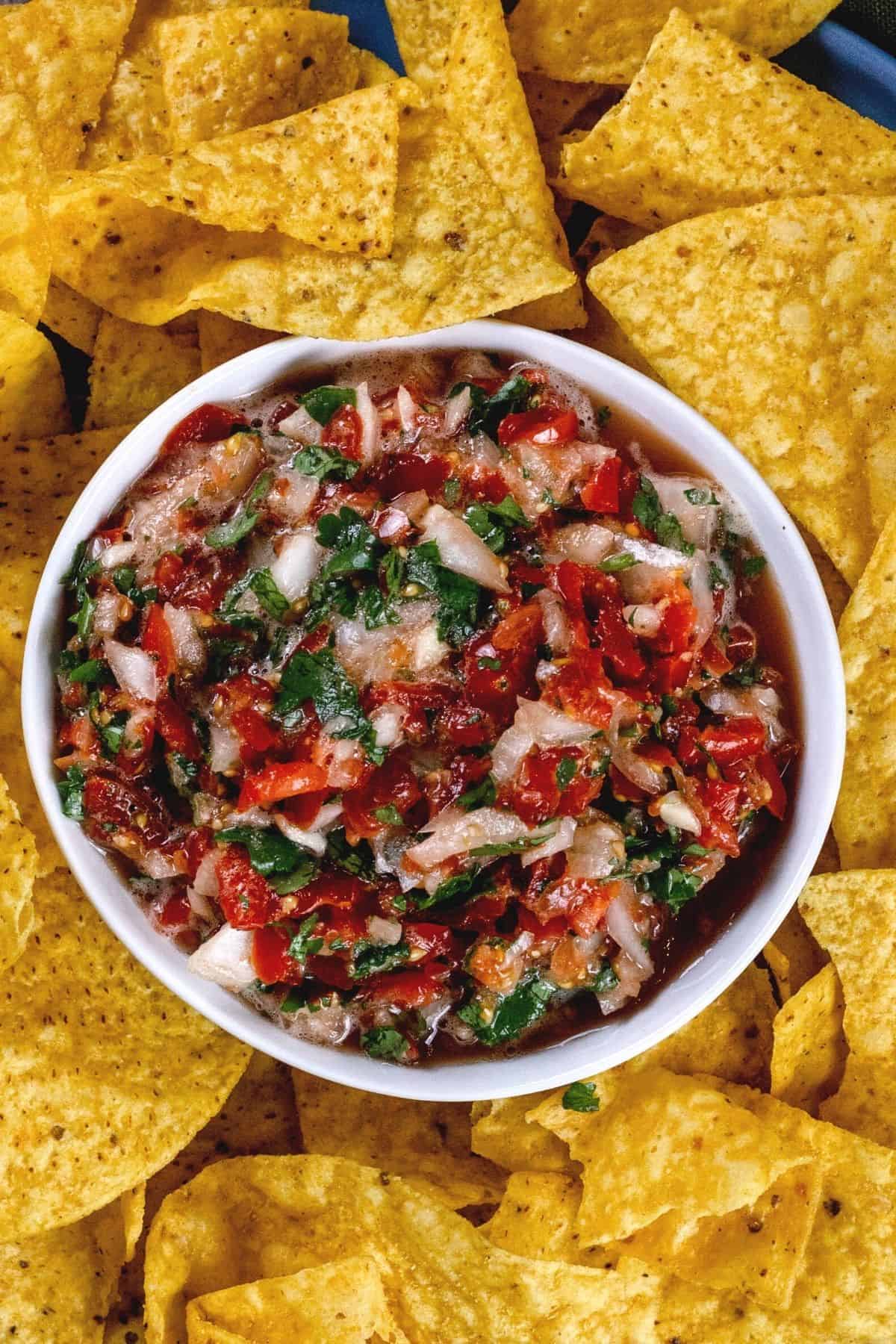 A white bowl filled with mild Pico de Gallo that is surrounded by yellow corn tortilla chips.