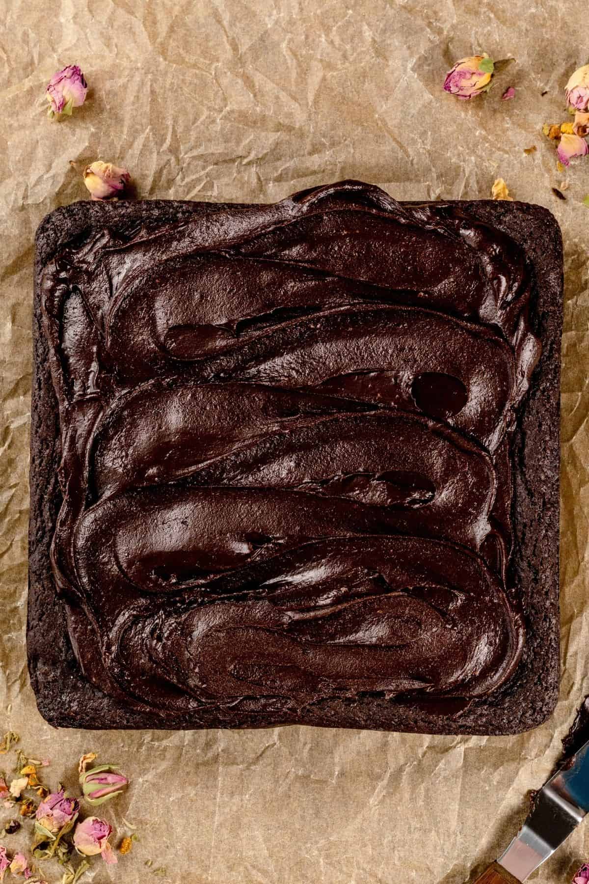 A batch of brownies on crinkled parchment paper with rose petals scattered around. An offset spatula is in the lower right corner. Thick chocolate frosting covers the brownies.