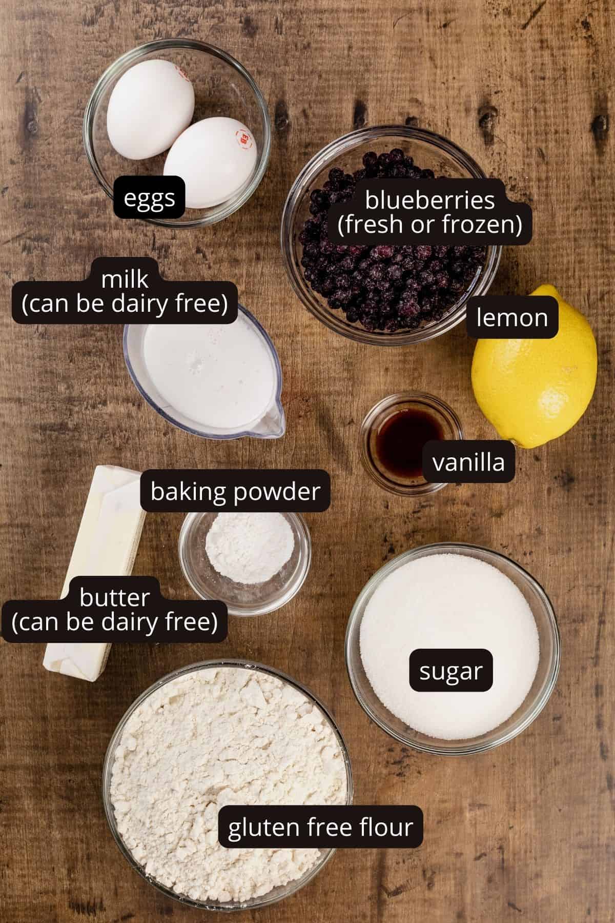 Ingredients for gluten free blueberry muffins in various glass bowls on a wood table. Black and white labels have been added to name each ingredient.
