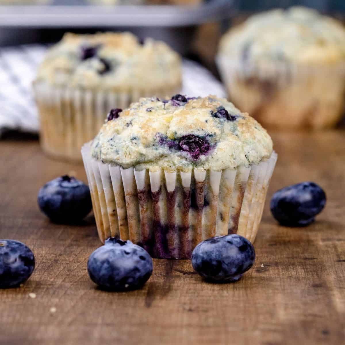 A gluten free blueberry muffin in a white muffin wrapper is next to fresh blueberries on a wood table. More muffins are blurred in the background.
