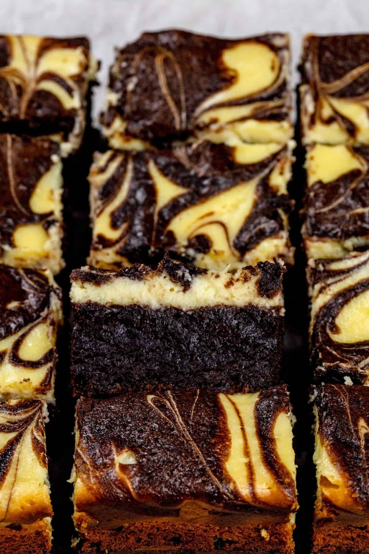 Looking down at the many slices of brownie in rows on white parchment paper. One brownie is on it's side so you can see the layers of brownie, cheesecake, and swirls.