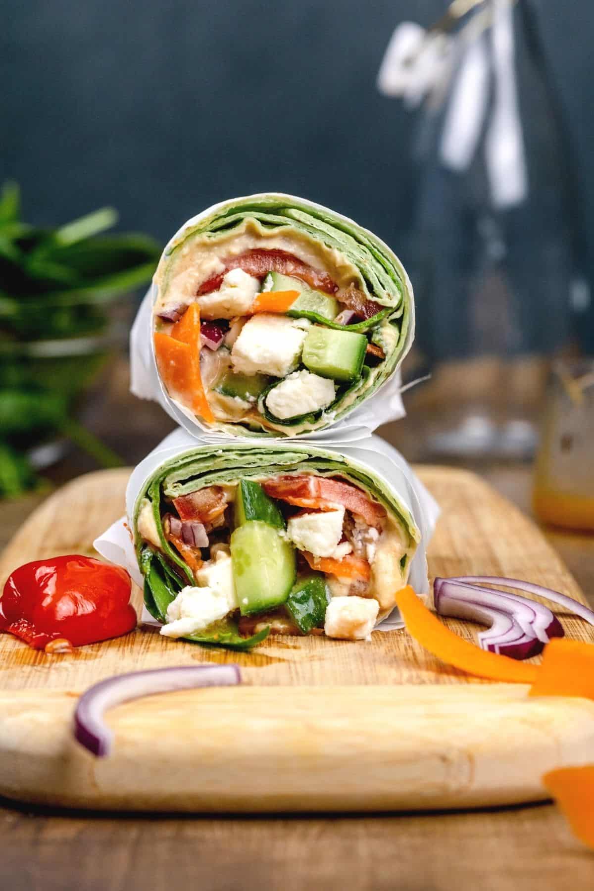 A stacked veggie wrap with veggies overflowing in the wrap and onto the wood cutting board. More veggies and a glass bottle are blurred in the background.