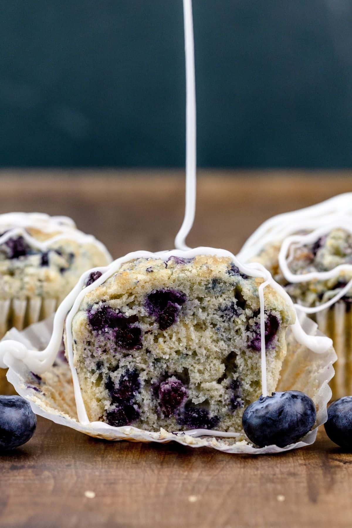 3 blueberry muffins on a wood table with fresh blueberries. The muffin in front has been cut in half so you can see the texture and crumb of the muffin. A white drizzle of icing is slowly being poured over the muffins.