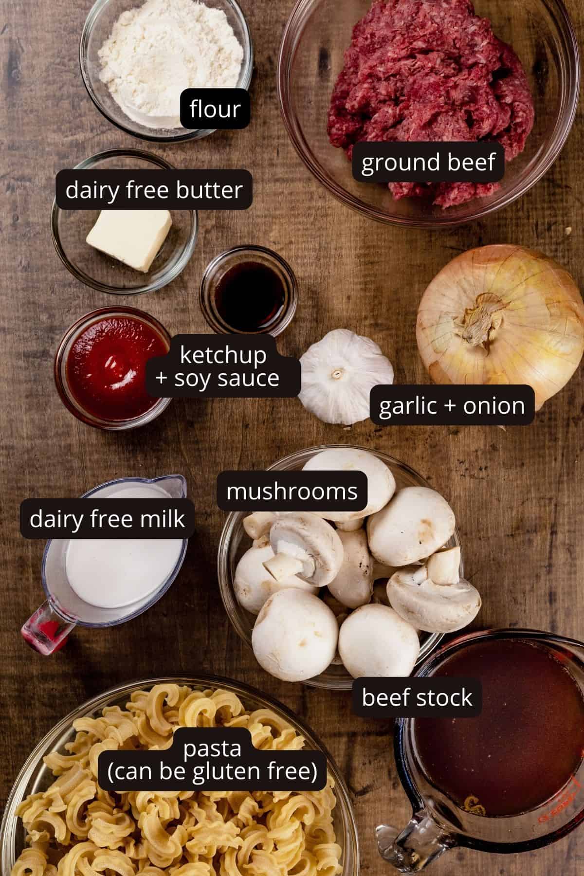 Ingredients for dairy free beef stroganoff in various glass bowls on a wood table top. Black and white labels have been added to name each ingredient.