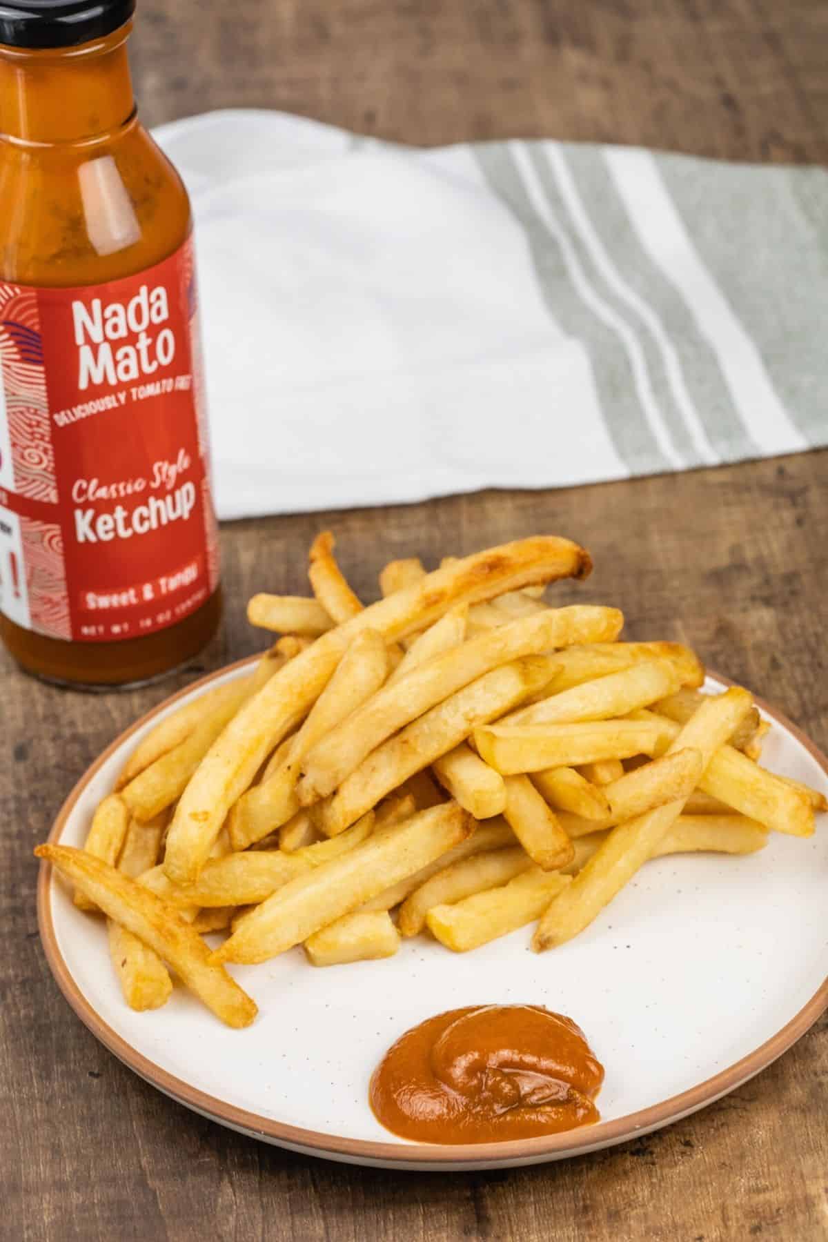 A small beige plate with French fries and a big dollop of Nada Mato ketchup on the plate. The bottle is behind the plate. A green and white towel is blurred in the background.