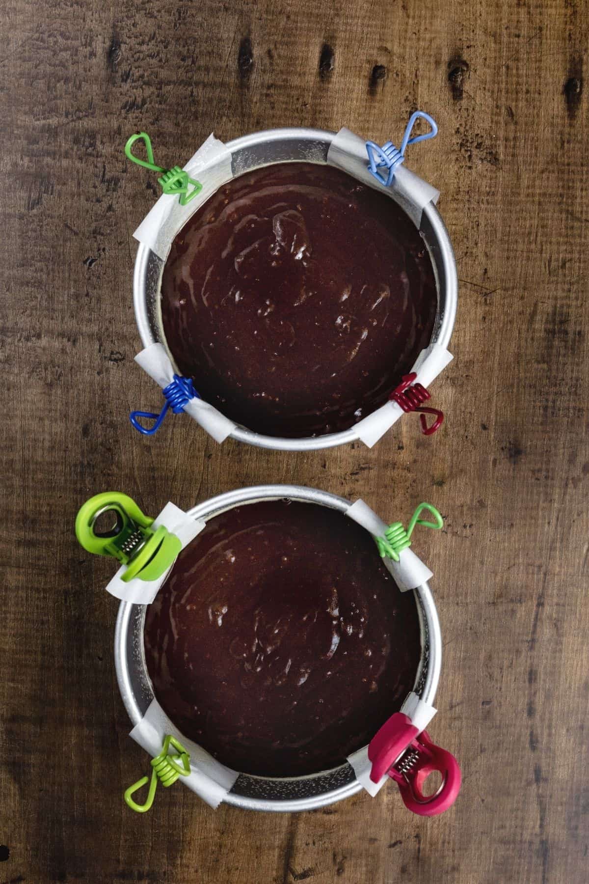 Two 6-inch cake pans lined with parchment and filled with raw cake batter. Little clips have been added to help hold the parchment in place.