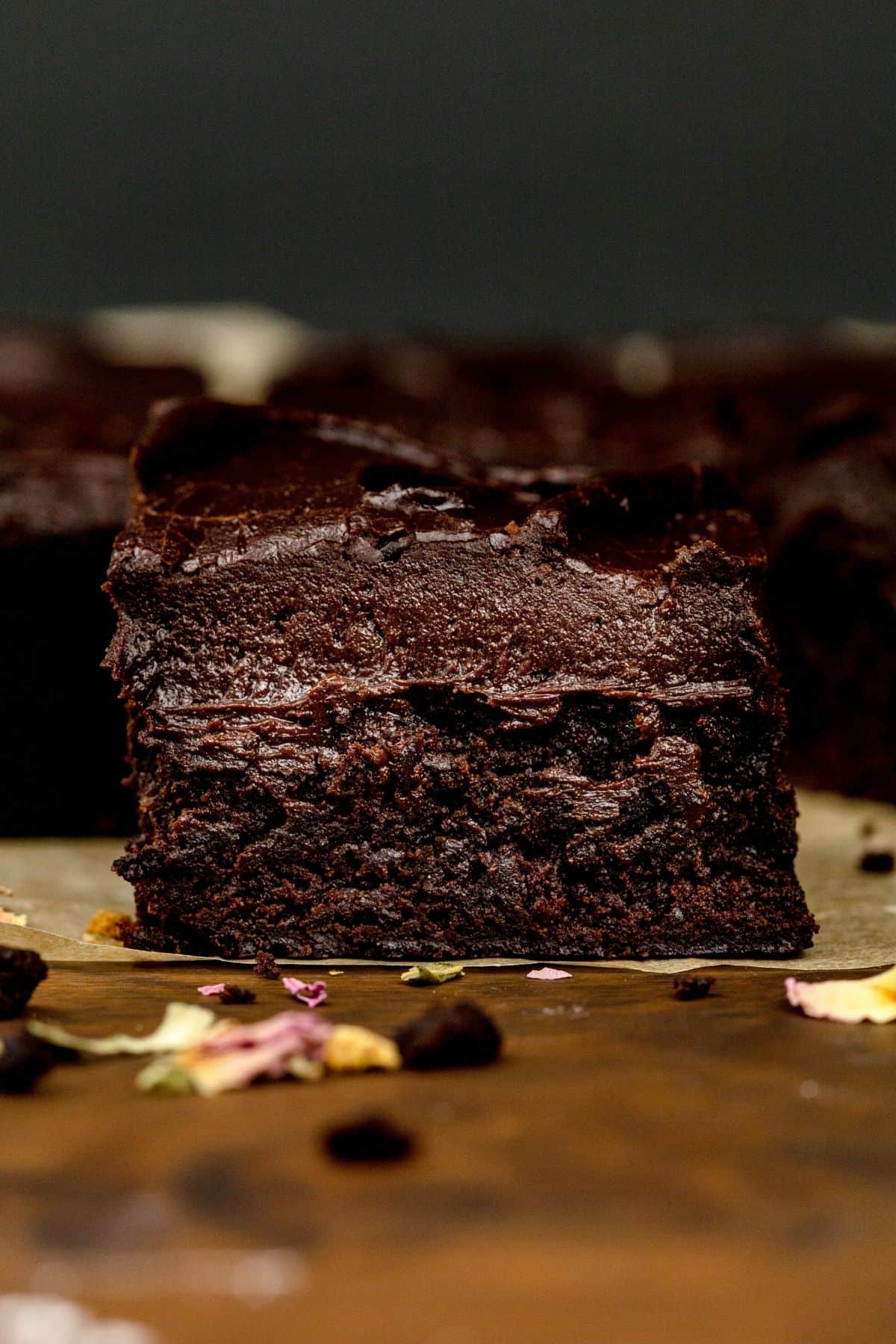 A side view of a single brownie covered in thick chocolate frosting. Rose petals and parchment paper are seen scattered around.