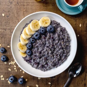White bowl filled with blueberry oatmeal and sliced bananas and more fresh blueberries on top. A few blueberries and oats are on the wood table next to the bowl. A small cup of coffee is in the top right corner.