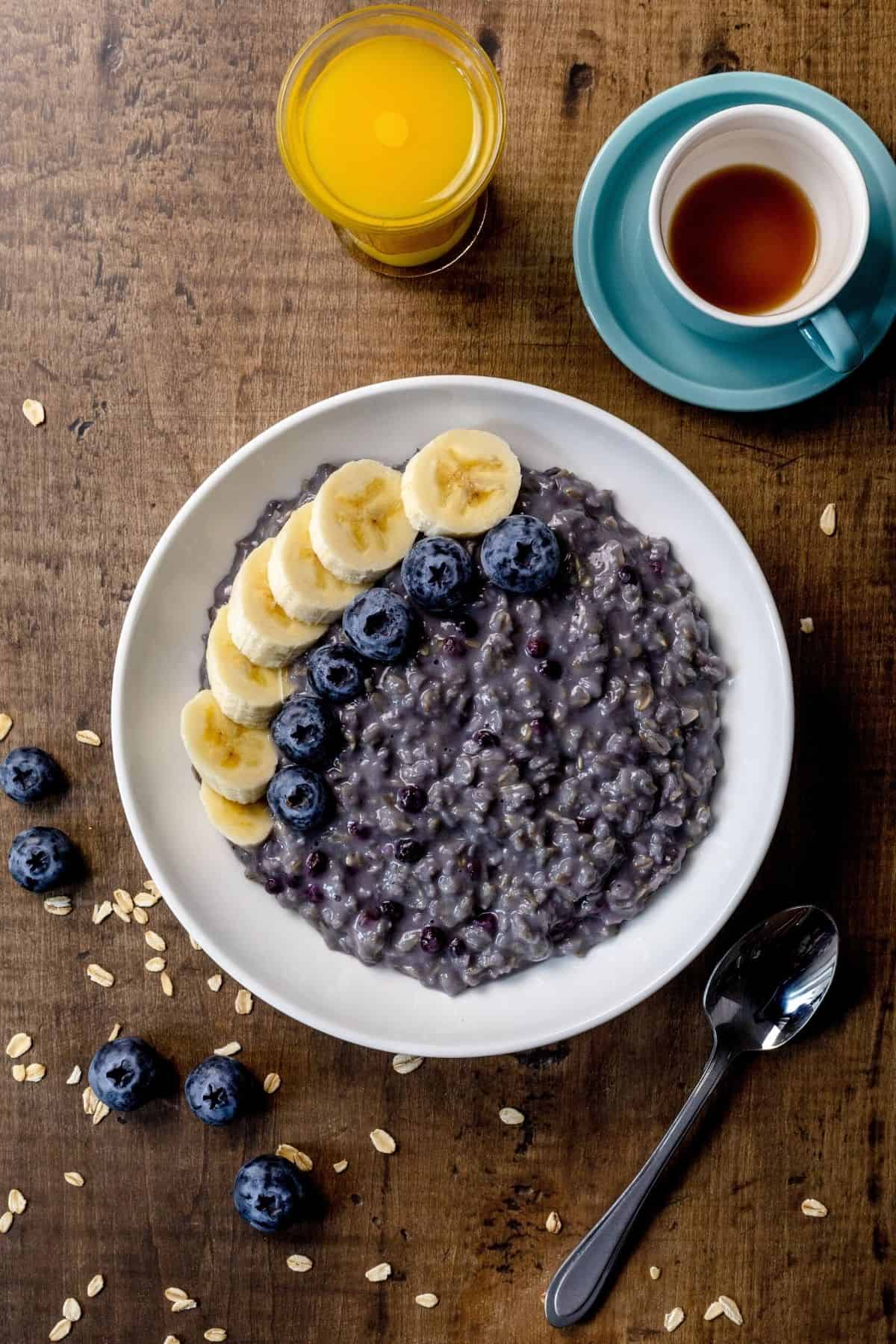 A single bowl of blueberry oatmeal on a wood tabletop. Sliced bananas and fresh blueberries are on top of the oatmeal. A spoon is next to the bowl. Fresh blueberries and oats are sprinkled around. A small cup of coffee and a small glass of orange juice are in the top right corner of the photo.