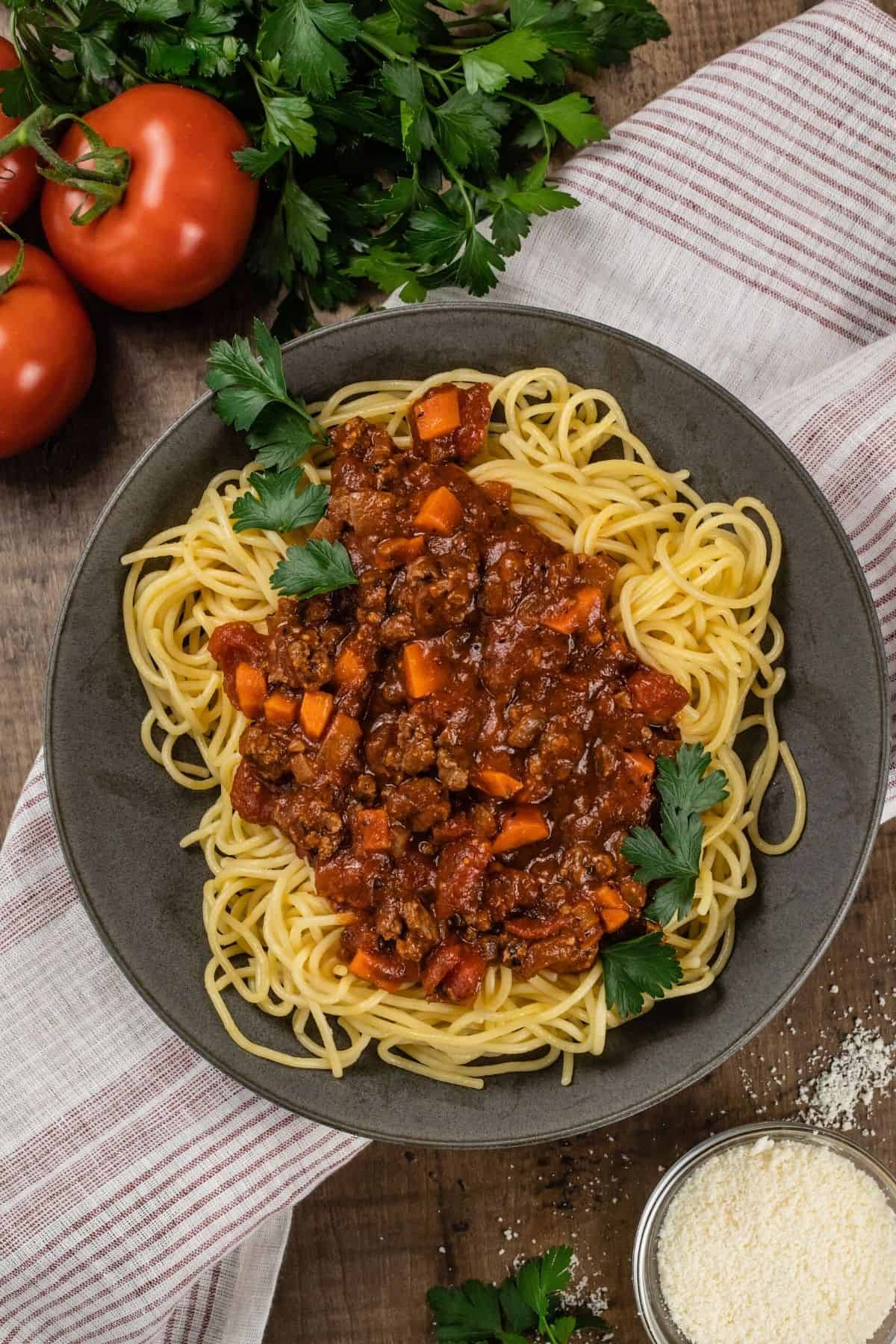A big bowl filled with spaghetti and beef bolognese sauce is sprinkled with fresh parsley. More parsley, tomatoes, and a small bowl of parmesan cheese are next to the bowl as it rests on a red and white towel on a wood table.