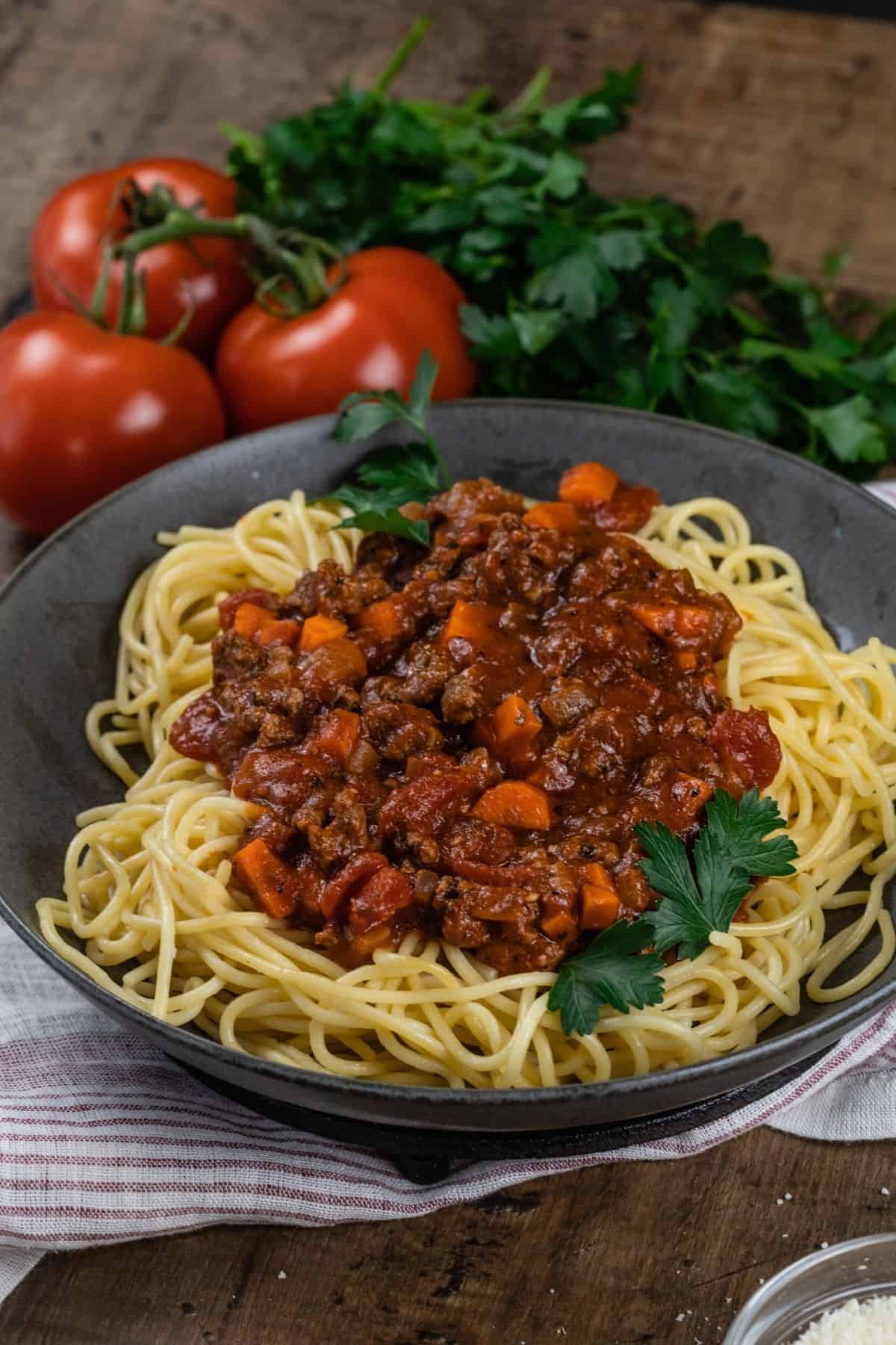 Side view of a big bowl filled with spaghetti and beef bolognese sauce is sprinkled with fresh parsley. More parsley, tomatoes, and a small bowl of parmesan cheese are next to the bowl as it rests on a red and white towel on a wood table.