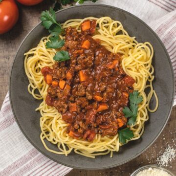 A big bowl filled with beef bolognese sauce and spaghetti noodles. Fresh parsley is sprinkled on top. Fresh tomatoes and a tiny bowl of parmesan cheese are in the corners.