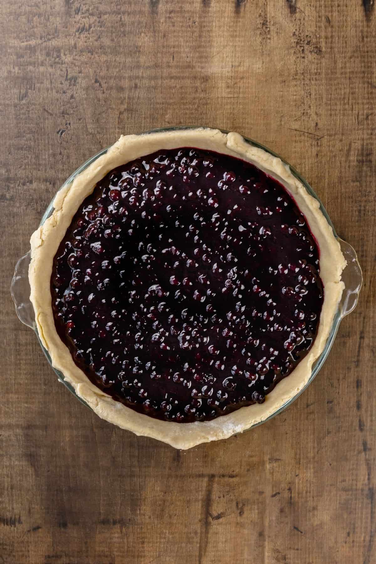 An unbaked blueberry pie with the finished filling in a glass pan on a wood table.