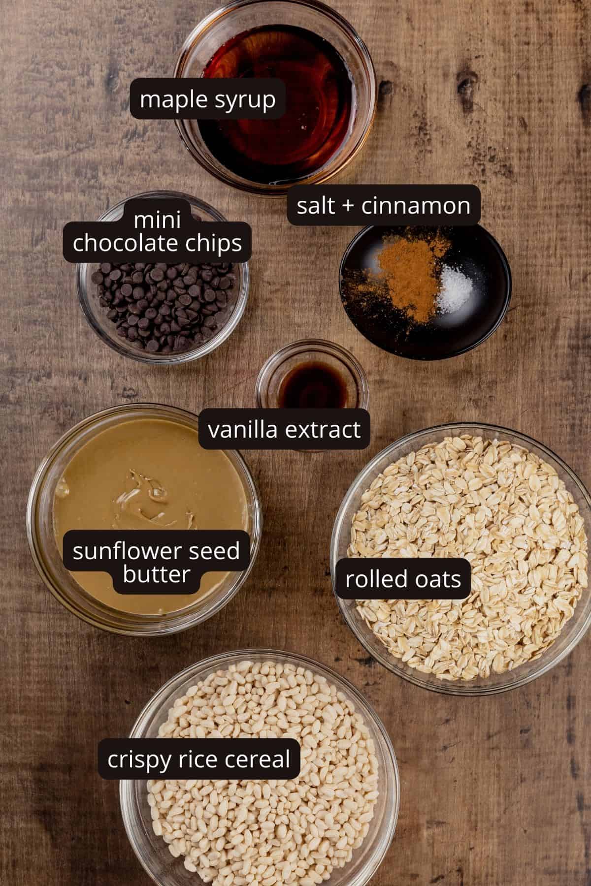 Ingredients for nut free granola bars in various bowls are on a wood tabletop. Black and white labels have been added to name each ingredient.