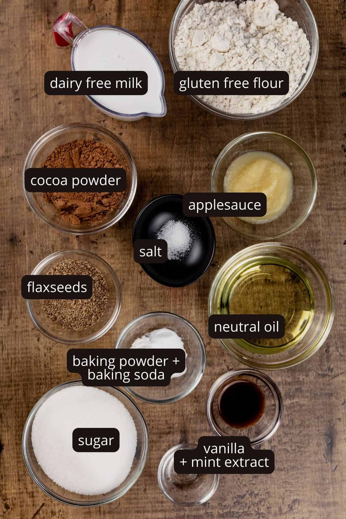 Ingredients for mint chocolate cupcakes in various glass bowls on the wood kitchen table. Black and white labels have been added to name each ingredient.