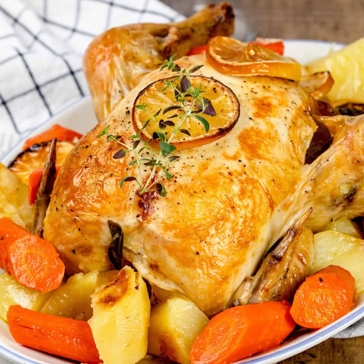 Close up of a whole lemon garlic roasted chicken in a serving dish with roasted carrots and potatoes.