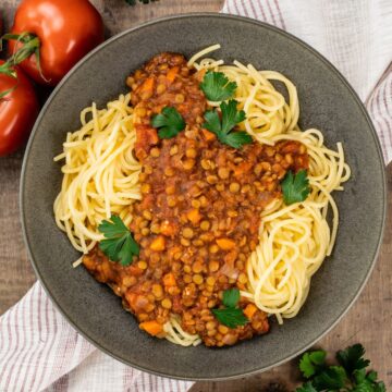 Big bowl filled with spaghetti pasta and covered in a lentil bolognese sauce. Bits of fresh parsley dot the dish. More parsley and fresh tomatoes are next to the bowl. The bowl is on a red and white stripe towel on a wood table.