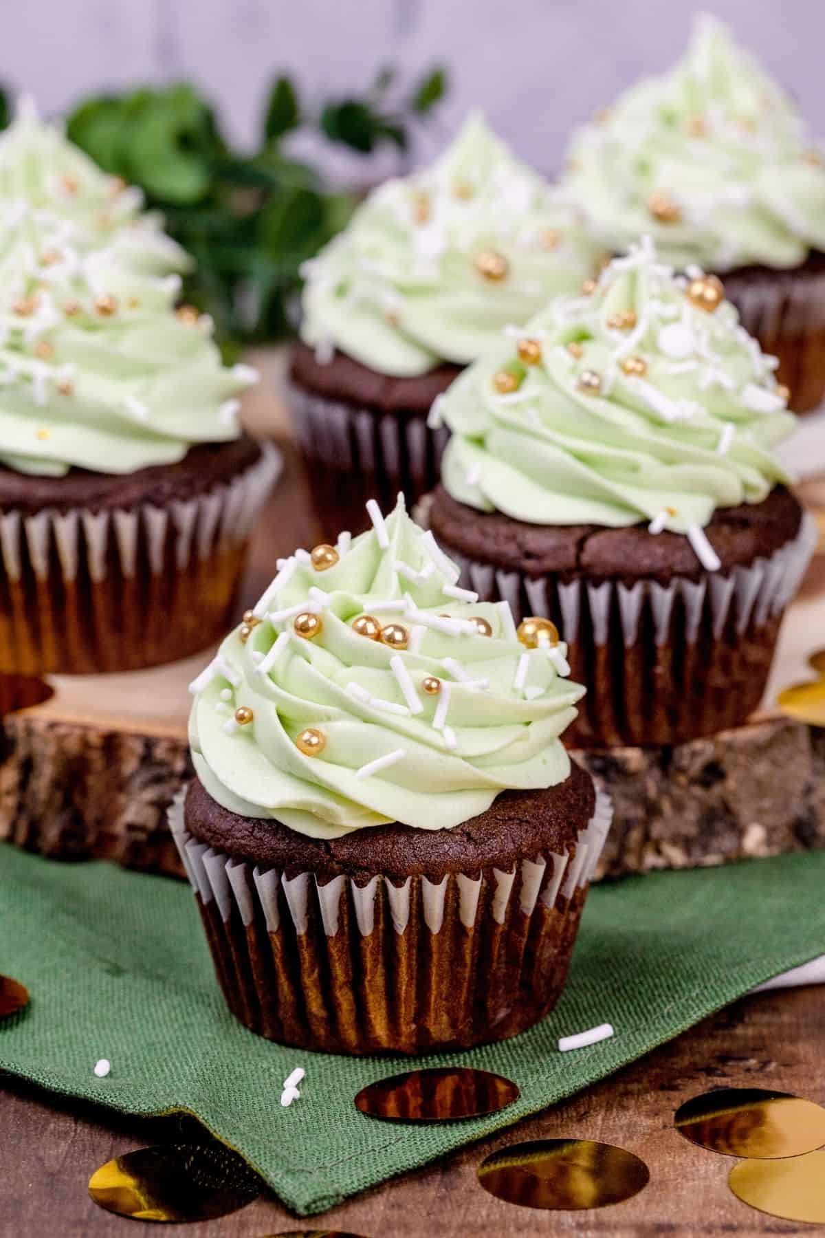 Many chocolate cupcakes with green mint frosting and gold and white sprinkles. One is on a green napkin and more are on a sliced wood platter. Sprinkles and gold foil confetti are scattered around the scene.